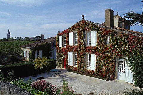Ein malerisches Landhaus mit roten und grünen Ranken bedeckt. Vor dem Haus befindet sich ein kleiner Außenbereich mit einem Tisch und Stühlen. Im Hintergrund ist ein Kirchturm sichtbar, umgeben von Weinbergen.
