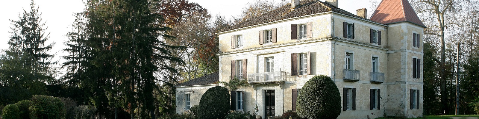 Ein elegantes, mehrstöckiges Landhaus mit einem spitzen Dach turm steht in einer grünen Umgebung, umgeben von hohen Bäumen und gepflegten Büschen. Es hat große Fenster mit Holzläden.