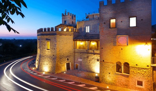Eine beeindruckende Burg steht am Straßenrand, beleuchtet von warmem Licht. Der Abendhimmel fängt an zu dämmern, während Fahrbahnlichter sanft um die Kurve fließen.