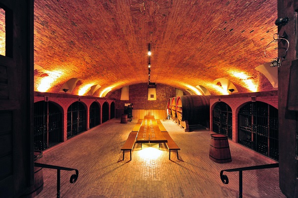 Ein Weinkeller mit einer langen Holztischreihe in der Mitte. An den Wänden stehen große Fässer und Regale mit Weinflaschen. Die Decke ist gewölbt und aus Ziegeln, sanft beleuchtet.