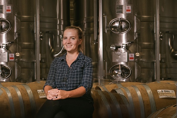 Eine lächelnde Frau sitzt auf einem Holzfass in einer Brauerei, umgeben von großen silbernen Tanks. Sie trägt ein kariertes Hemd und hat die Haare zurückgebunden.