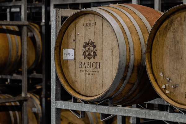 Ein spezielles Holzfass mit der Aufschrift -BABICH- und einem Wappen aus New Zealand Wine steht auf einem Metallregal in einem Lagerraum, umgeben von weiteren Fässern.