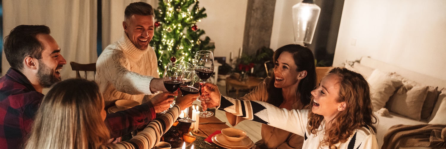 Eine Gruppe von sechs Personen hebt Gläser mit Rotwein an, während sie an einem festlich gedeckten Tisch sitzen. Im Hintergrund steht ein beleuchteter Weihnachtsbaum. Gemütliche Atmosphäre.