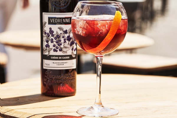 Ein elegantes Cocktailglas steht auf einem Holztablett, gefüllt mit einem rot gefärbten Getränk und Eiswürfeln, garniert mit einer Orangenscheibe. Im Hintergrund ist eine Flasche Crème de Cassis sichtbar.