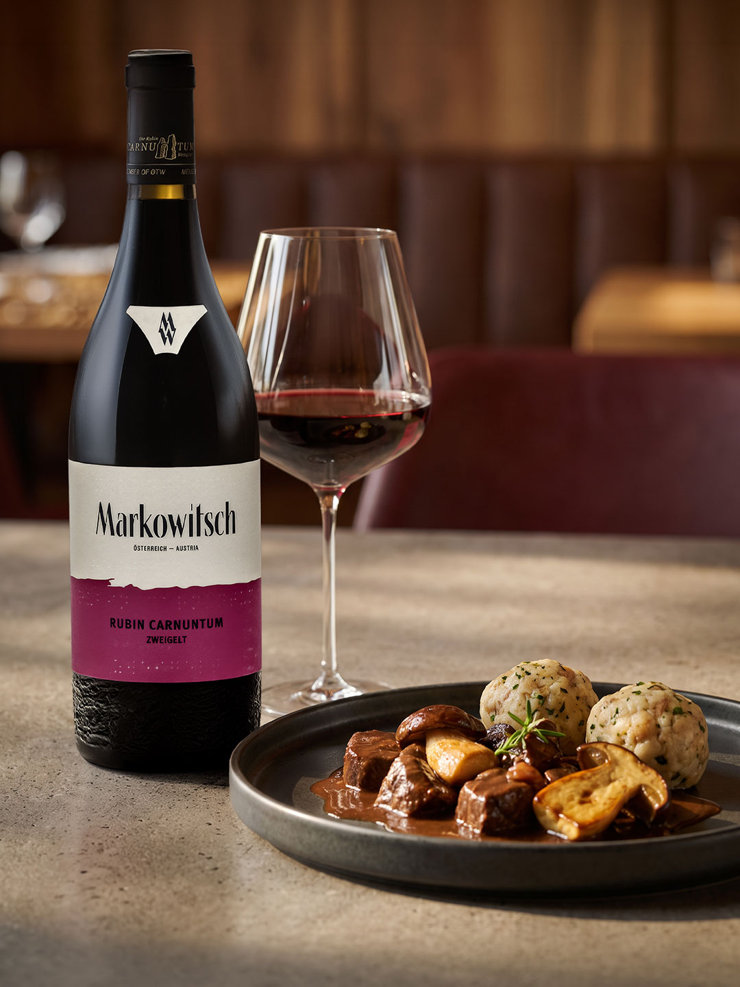 Eine Flasche Rotwein steht neben einem Weinglas, während ein Teller mit Fleischstücken, Pilzen und Knödeln auf einem Tisch präsentiert wird. Der Hintergrund zeigt eine gemütliche Restaurantatmosphäre.