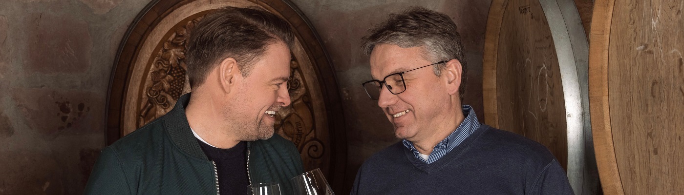 GIER auf Wein Zwei Männer stehen in einem Weinkeller und lachen sich an, während sie Gläser in der Hand halten. Im Hintergrund sind große Holzfässer zu sehen, die die typische Atmosphäre eines Weinkellers vermitteln.