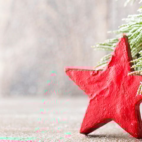Ein roter Stern aus Holz steht auf einer neutralen Oberfläche, umgeben von grünen Tannenzweigen. Die Szene hat eine weihnachtliche Stimmung.