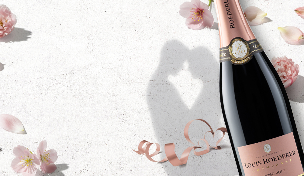 Eine Flasche Louis Roederer Rosé Champagner steht vor einer hellen, strukturierten Wand. Rundherum sind zarte Blüten und dekorative, spiralförmige Bänder angeordnet. Im Hintergrund ist eine Schattenumriss von zwei Personen, die sich küssen, zu sehen.