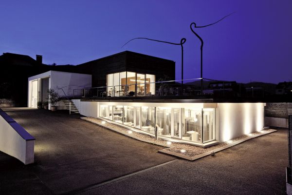 Modernes Gebäude mit großen Fensterfronten, beleuchtet von unten, in einer ruhigen Umgebung. Ein kreatives, skulpturales Element erhebt sich darüber. Dämmerungsstimmung mit tiefblauem Himmel.
