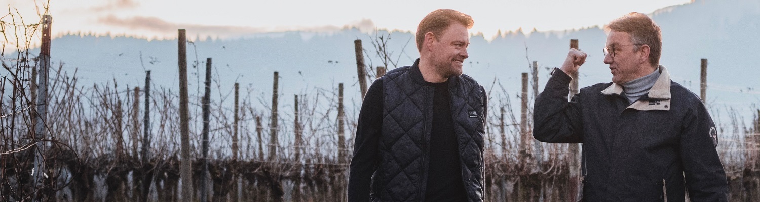 Auf einen Wein mit...Max Giermann Zwei Männer gehen lächelnd durch die Weinreben, umgeben von sanften Hügeln im Hintergrund. Es ist wahrscheinlich Morgen oder Abend, da das Licht weich und warm ist.
