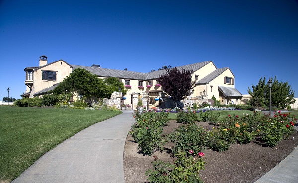 Ein großes, elegantes Gebäude mit einem gewölbten Dach steht im Hintergrund. Ein gepflasterter Weg führt durch grüne Rasenflächen und blühende Rosenbeete. Die Umgebung ist klar und sonnig.