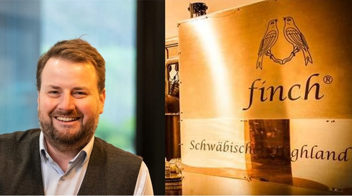 Ein lächelnder Mann mit Bart trägt eine Weste und sitzt neben einem goldenen Schild mit dem Wort -finch- und dem Schriftzug -Schwäbische Highland-.