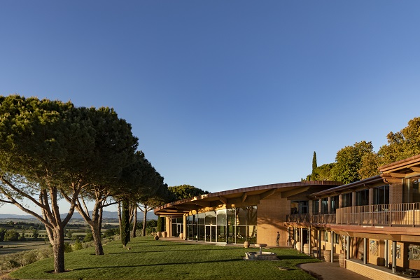 Ein modernes Gebäude mit vielen Fenstern steht auf einer grünen Wiese, umgeben von großen Bäumen. Der Himmel ist klar und blau, die Landschaft erstreckt sich im Hintergrund.