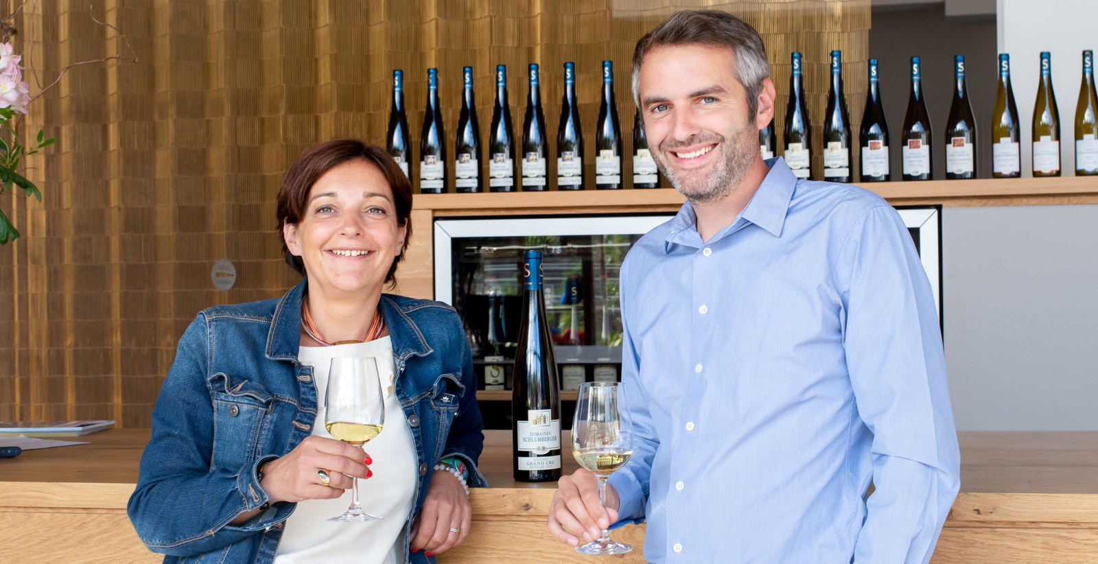 Zwei Personen stehen an einer Holztheke und halten jeweils ein Weinglas in der Hand, während im Hintergrund mehrere Weinflaschen aufregend angeordnet sind. Sie lächeln freundlich.