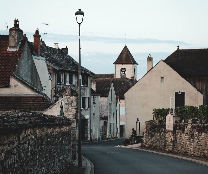 Eine enge, geschwungene Straße in einem kleinen Dorf mit historischen Gebäuden, einer Straßenlaterne und einem Kirchturm im Hintergrund, unter einem bewölkten Himmel.