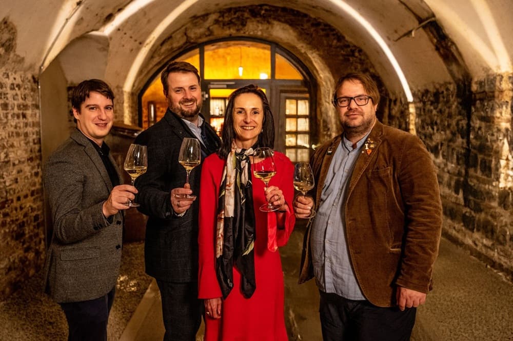 Vier Personen stehen in einem Keller mit Steinwänden, halten Weingläser und lächeln. Eine Frau in einem roten Kleid steht in der Mitte, während die anderen drei Herren nebeneinander stehen.