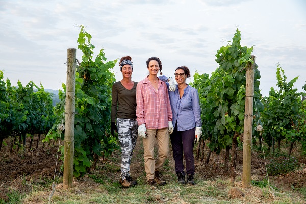 Drei Frauen stehen lächelnd zwischen Weinreben in einem Weinberg. Sie tragen praktische Kleidung und Gärtnerhandschuhe. Der Himmel ist teilweise bewölkt, und die Umgebung ist grün und ländlich.