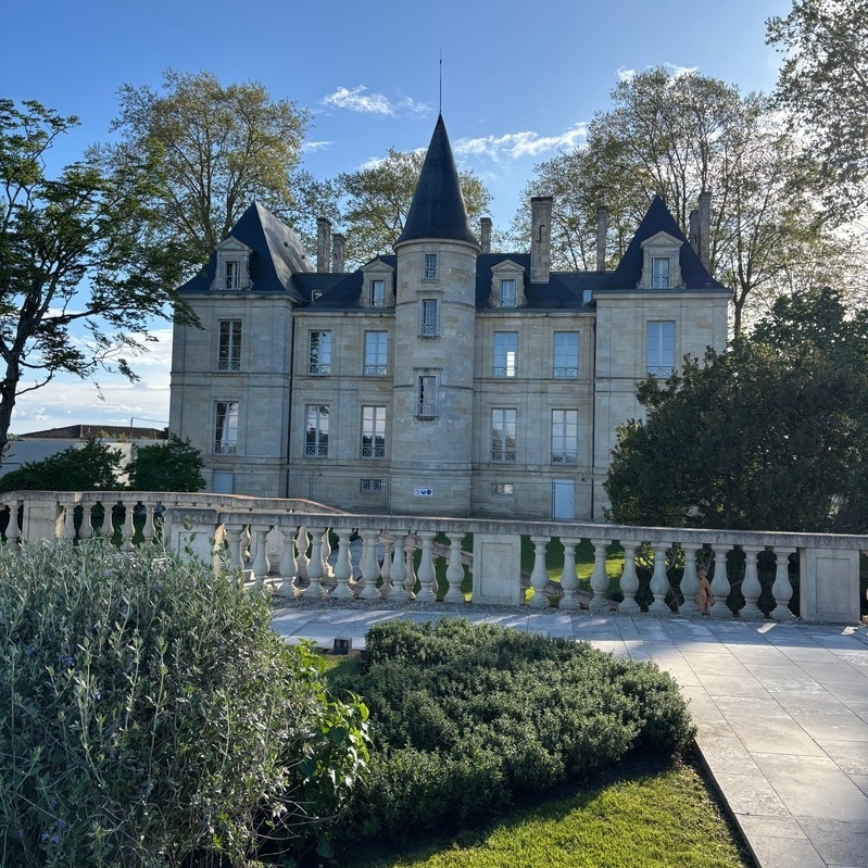 Ein historisches Schloss mit einem Spitzdach und mehreren Fensterreihen steht im Mittelpunkt. Es ist von einem gepflegten Garten mit Büschen und einem eleganten Geländer umgeben, unter blauen Himmel.