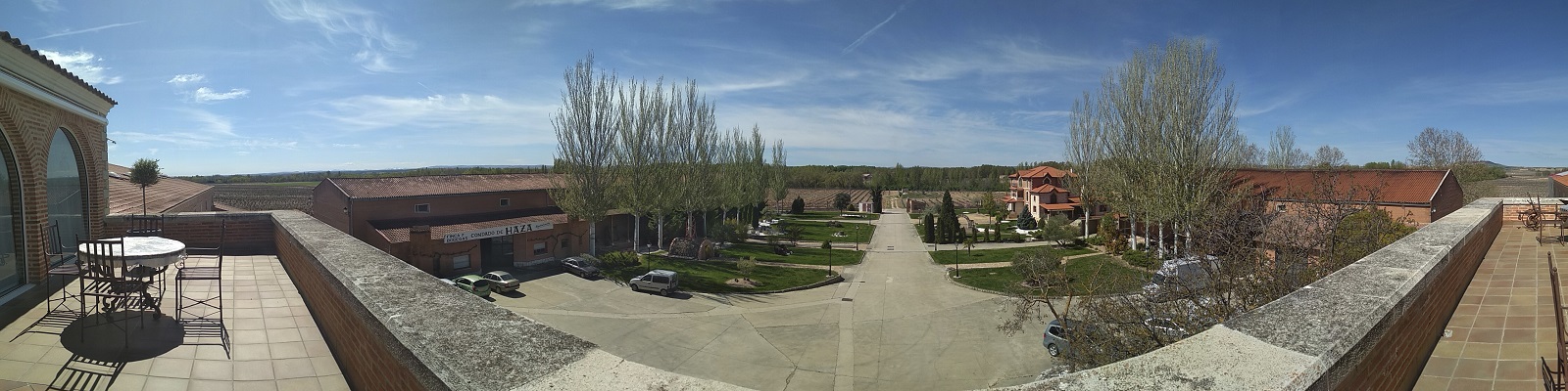 Condado de Haza Ein Balkon mit einem runden Tisch und Stühlen blickt auf eine weitläufige Anlage mit gepflasterten Wegen, Gebäuden und Bäumen unter einem klaren blauen Himmel.