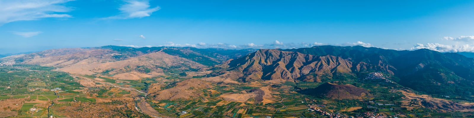 Ein großer Berg dominiert die Landschaft, umgeben von üppigen, grünen Feldern und vereinzelten Häusern. Der Himmel ist leicht bewölkt, was die ruhige Atmosphäre unterstreicht.