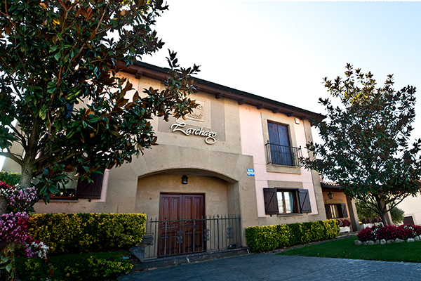 Ein zweistöckiges Gebäude mit einem holzverkleideten Eingang und Balkonen. Es ist von grünen Büschen und Bäumen umgeben, und der Schriftzug -Zarchago- ist deutlich sichtbar.