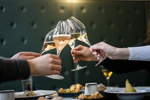 Fünf Gläser mit Sekt stoßen zusammen, während Hände von verschiedenen Personen auf einem Tisch mit elegantem Essen und einem grünen Hintergrund sichtbar sind.