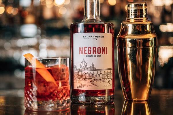 Eine Flasche Negroni mit einem Etikett sitzt neben einem Cocktailshaker und einem Glas mit einem Negroni-Cocktail, garniert mit einer Orangenscheibe, in einer stilvollen Bar-Umgebung.