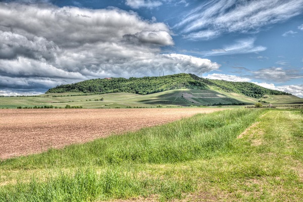 Eine hügelige Landschaft mit grasbewachsenen Feldern. Im Vordergrund sind braune Ackerflächen, während im Hintergrund grüne Hügel unter einem bewölkten, blauen Himmel sichtbar sind.