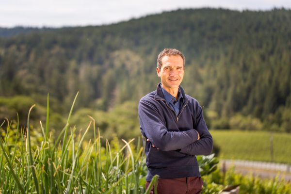 Ein Mann steht mit verschränkten Armen in einem Feld mit grünen Pflanzen. Im Hintergrund erstrecken sich bewaldete Hügel und eine Weinberg-Landschaft unter einem bewölkten Himmel.