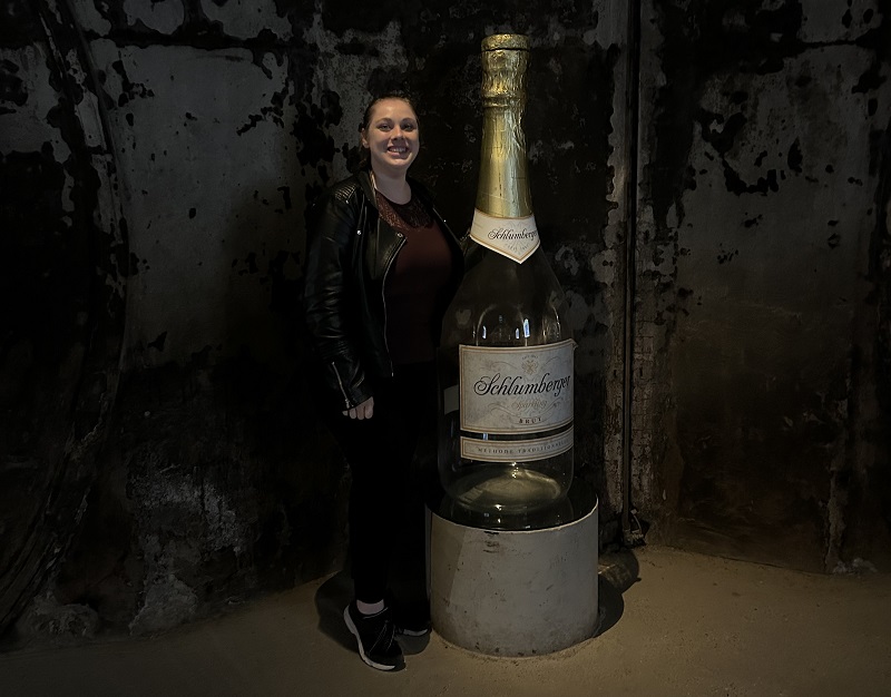 Eine Frau steht neben einer riesigen Sektflasche mit dem Label -Schlumberger-. Der Hintergrund ist eine roh wirkende Wand in einem dunklen Raum. Die Frau lächelt und trägt eine schwarze Lederjacke.