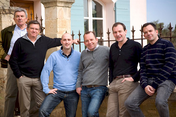 Sechs Männer stehen lässig vor einem Gebäude mit einem hellen Außenanstrich und blauen Fensterläden. Sie posieren in entspannter Haltung, einige haben die Hände in den Taschen.