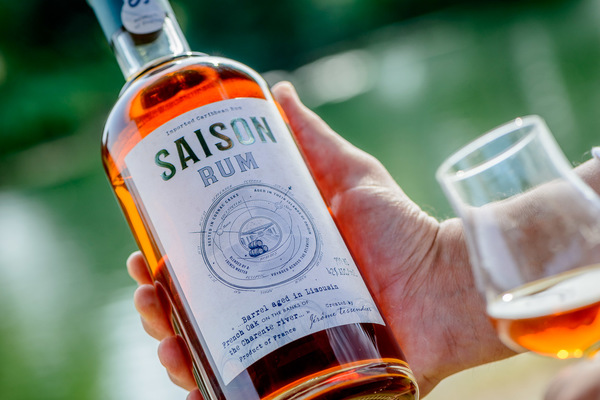 Eine Person hält eine Flasche Saison Rum in der Hand. Der Rum hat eine bernsteinfarbene Flüssigkeit und das Etikett ist gut sichtbar. Im Hintergrund sind unscharfe grüne Natur-Elemente zu erkennen.