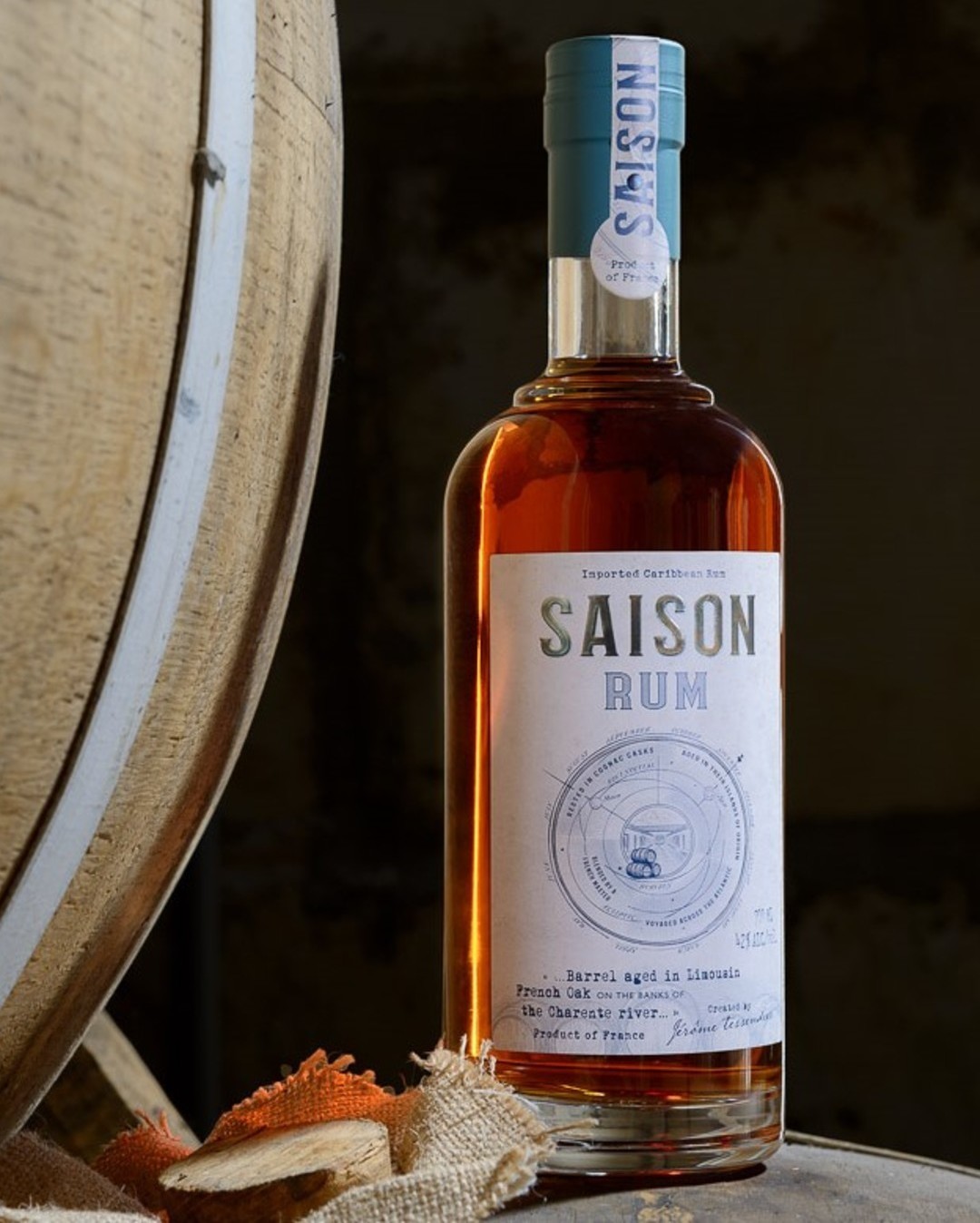 Eine Flasche Saison Rum steht vor einem Holzfass. Die Flasche hat ein helles Etikett mit Informationen über den Rum, einschließlich der Herkunft und Alterung. Die Umgebung ist rustikal und dunkel.