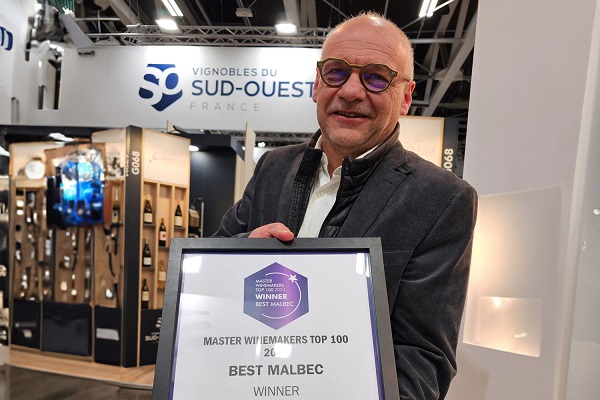 1_31.07._Bertrand-Vigouroux-Master-WineMakers-Top-100-Best-Malbec-Winner - 600 Ein Mann hält eine Urkunde mit der Aufschrift -Best Malbec Winner- in einer Weinmesse-Umgebung, die mit Weinflaschen und einem Banner mit dem Namen -Vignobles du Sud-Ouest, France- dekoriert ist.