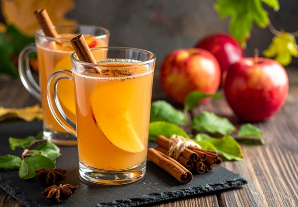 Heiße Getränke in transparenten Gläsern stehen auf einem dunklen Untergrund, umgeben von Äpfeln und grünen Blättern. Zimtstangen sind in den Gläsern und daneben arrangiert.
