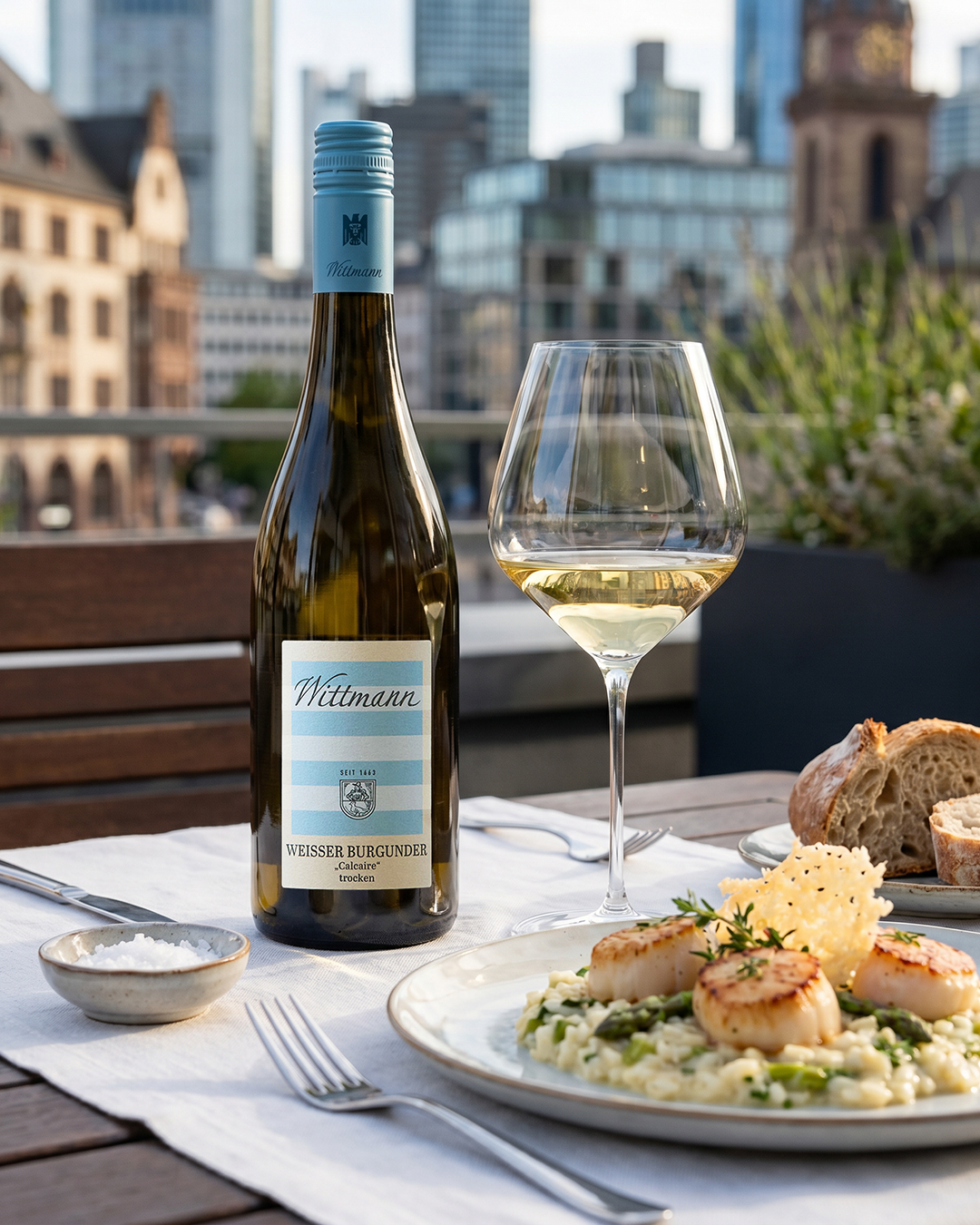 Eine Flasche Weißer Burgunder steht neben einem Glas Wein auf einem Tisch. Daneben liegt ein Teller mit Risotto und Jakobsmuscheln sowie ein Stück Brot. Im Hintergrund sind moderne Gebäude zu sehen.