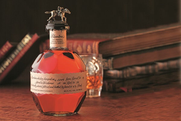 Ein Flakon mit rötlichem Whiskey steht auf einem Holztisch. Daneben befinden sich einige alte Bücher und ein Glas mit Whiskey. Oben auf dem Flakon befindet sich eine kleine Pferdefigur.