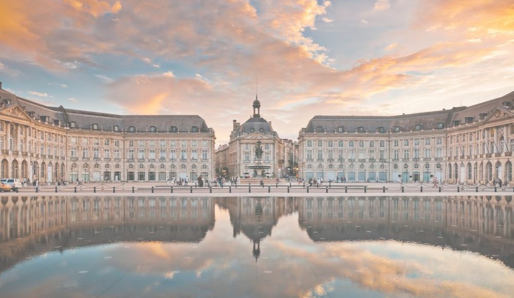 Eine große Platzlandschaft mit eleganten, historisch gestalteten Gebäuden umsäumt von Wasser, das die Architektur reflektiert. Der Himmel ist mit pastelartigen Wolken in warmen Farben gefärbt.