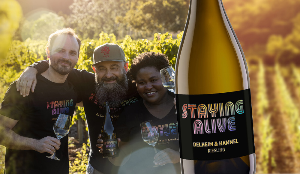 Drei Personen stehen in einem Weinberg, lächeln und halten Gläser in der Hand. Im Vordergrund steht eine Flasche Riesling mit dem Etikett -STAYING ALIVE- in bunten Buchstaben.