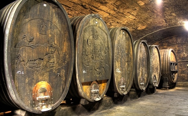 Fünf große, verzierte Fässer aus Holz stehen in einem unterirdischen Keller. Die Fässer sind mit reliefartigen Bildern geschmückt und in einem schwach beleuchteten Raum angeordnet.