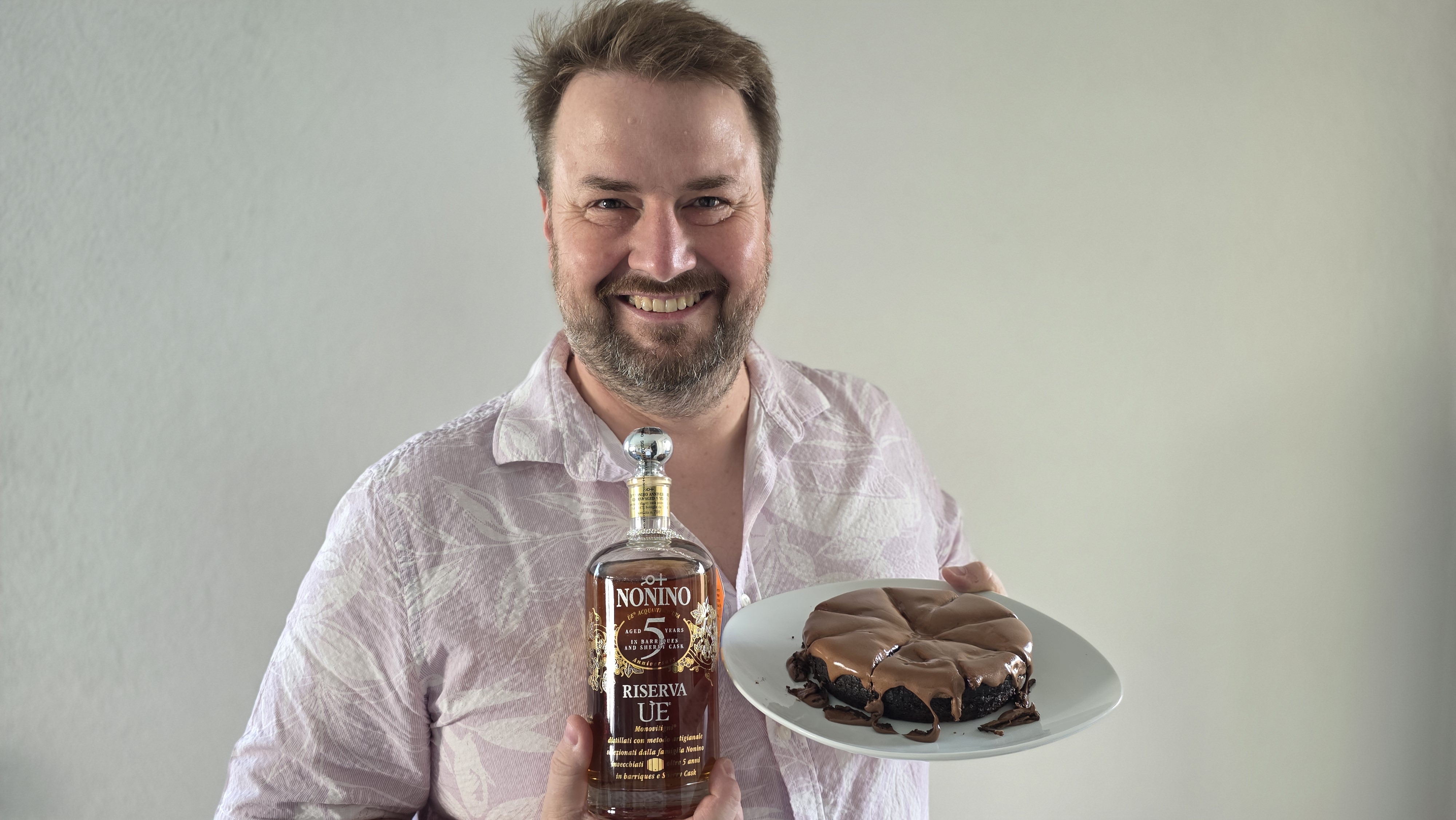 Ein Mann mit Bart lächelt und hält in der linken Hand eine Flasche Nonino Riserva UE und in der rechten ein Stück Schokoladenkuchen auf einem weißen Teller. Der Hintergrund ist einfarbig und hell.
