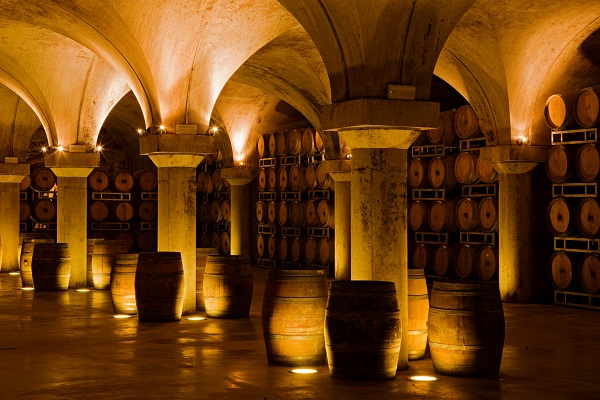 Holzfässer stehen auf dem Boden eines Weinkellers, umgeben von geschwungenen, beleuchteten Bögen und Regalen mit weiteren Fässern an der Wand, in einem gedämpften, warmen Licht.