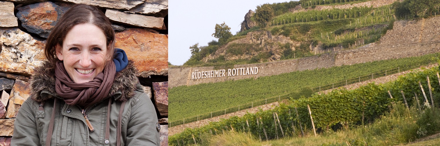 Auf einen Wein mit ... Theresa Breuer Eine Frau lächelt vor einer steinernen Wand, während im Hintergrund Weinberge mit dem Schriftzug -Rüdesheimer Rottland- sichtbar sind. Die Landschaft ist grün und hügelig, typisch für Weinbaugebiete.