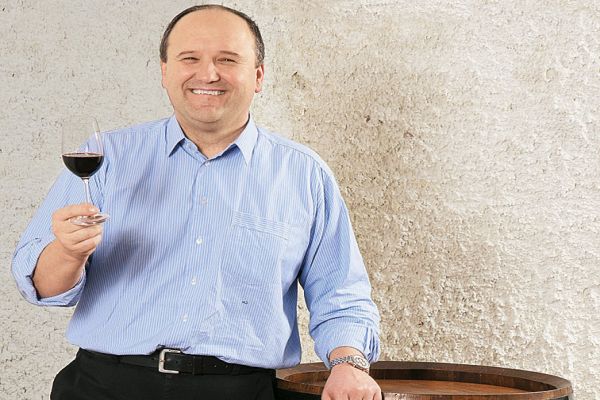 Ein Mann lächelt und hält ein Weinglas in der Hand. Er steht an einem Holzfass, vor einer wandähnlichen Struktur mit einem unregelmäßigen, hellen Muster.