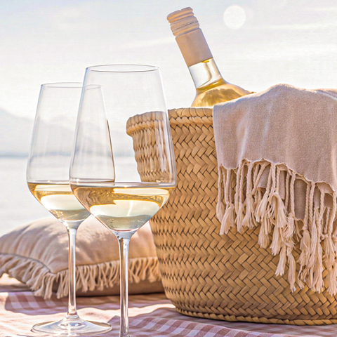 Zwei Weingläser mit weißem Wein stehen neben einem geflochtenen Korb, der eine Weinflasche und eine Decke enthält, auf einer Decke am Strand. Der Hintergrund zeigt eine ruhige Wasseroberfläche und sanfte Hügel.