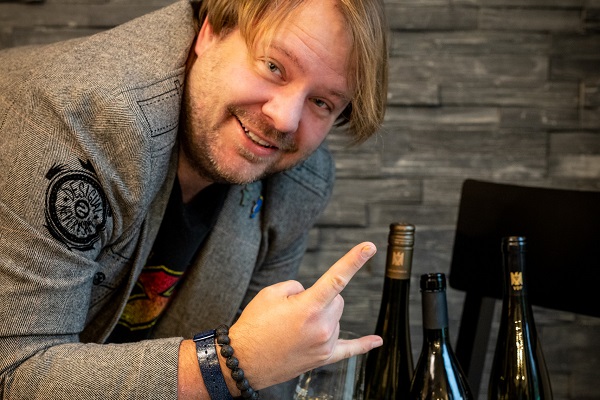 Ein Mann mit blonder Haarfrisur posiert fröhlich mit einem -Rock-on--Zeichen. Hinter ihm stehen mehrere Weinflaschen auf einem Tisch vor einer wandverkleideten Steinoberfläche.