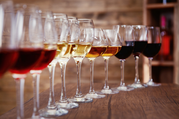 Eine Reihe von Weingläsern mit verschiedenen Weinsorten, darunter Rot-, Weiß- und Roséweine, steht auf einem Holztisch. Im Hintergrund sind Regale mit Getränken sichtbar.