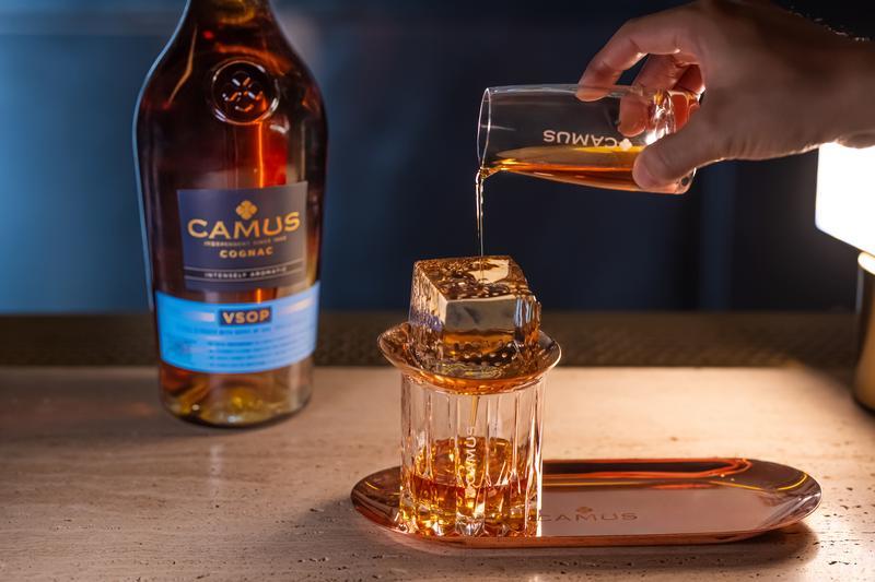Eine Hand gießt Cognac aus einem Glas über einen großen Eiswürfel in ein Glas mit Logo. Im Hintergrund steht eine Flasche Camus VSOP Cognac auf einem Tisch.