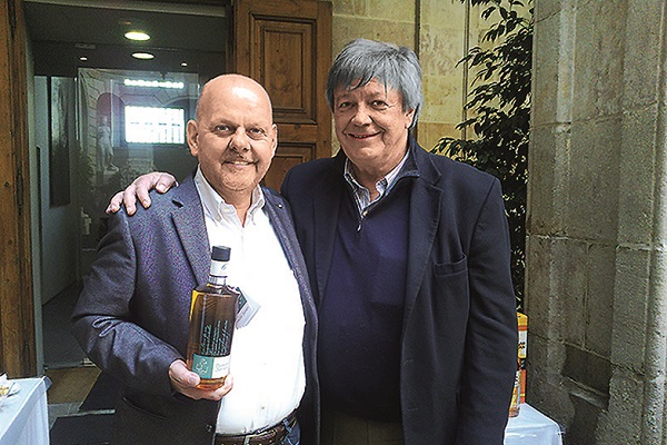 Zwei Männer stehen lächelnd nebeneinander und halten eine Flasche in der Hand. Sie sind in einem hellen Raum mit Holzdetails und Pflanzen im Hintergrund.