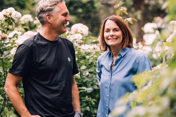 Zwei Personen stehen lächelnd in einem blühenden Garten voller Rosen. Sie scheinen sich zu unterhalten, während sie entspannt und freundlich wirken.
